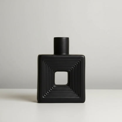 A matte black square bottle with a square cutout in the center and concentric ridged detailing on the front, shown on a light surface against a warm beige background. Small text in the upper left lists dimensions: four sides length 15.