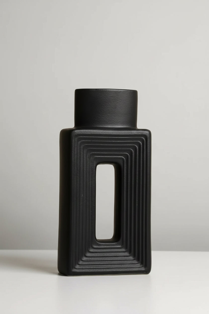 A matte black rectangular vase with a central cutout and concentric ribbed detailing, standing on a white surface against a beige background. Text in the upper left lists the dimensions.