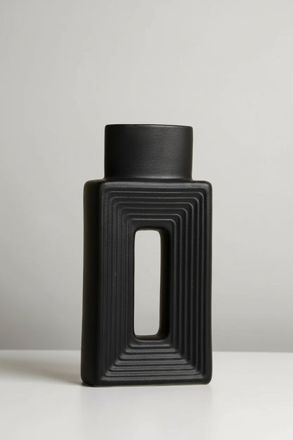 A matte black rectangular vase with a central cutout and concentric ribbed detailing, standing on a white surface against a beige background. Text in the upper left lists the dimensions.
