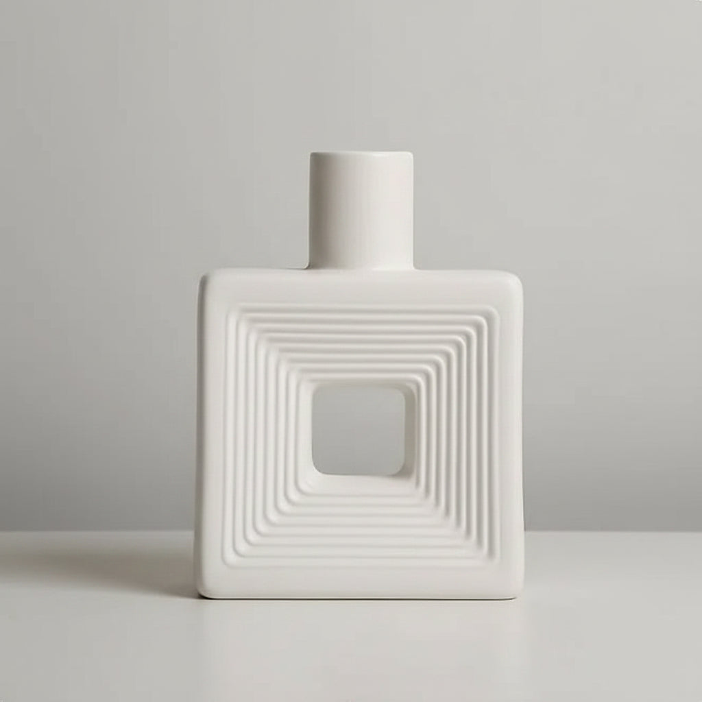 A white ceramic vase with a square body, concentric square ridges, and a central cutout, shown on a light surface against a warm beige background. Text in the top left lists dimensions.