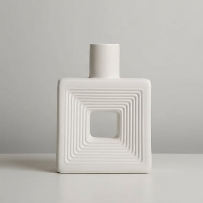 A white ceramic vase with a square body, concentric square ridges, and a central cutout, shown on a light surface against a warm beige background. Text in the top left lists dimensions.