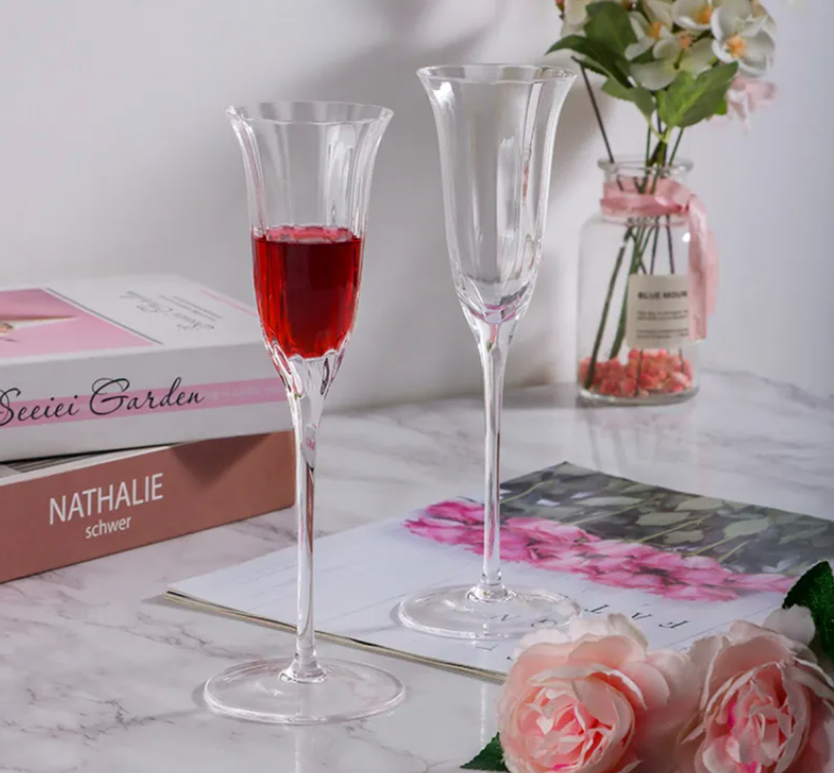 Two champagne flutes with red liquid on a marble surface with books and flowers in the background.