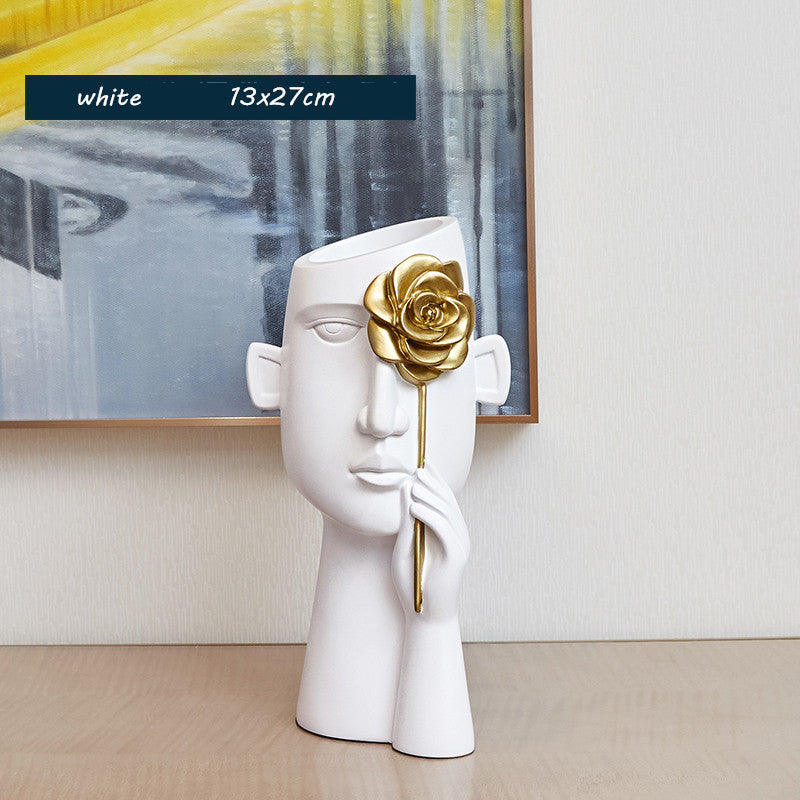 White ceramic sculpture with a gold flower held by a hand, placed on a wooden surface with an abstract painting in the background.