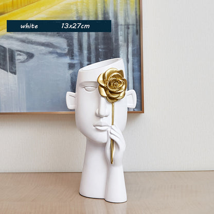 White ceramic sculpture with a gold flower held by a hand, placed on a wooden surface with an abstract painting in the background.
