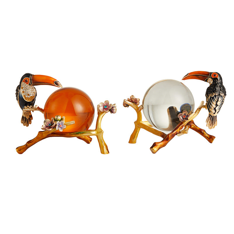 Two decorative items with toucan birds perched on branches holding glass balls on a white background.