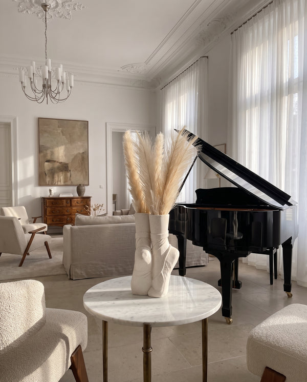 Elegant, sunlit living room featuring a grand piano, marble table with ballet shoe vase, and chic neutral decor.
